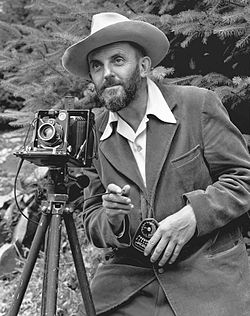A photo of a bearded Ansel Adams with a camera on a tripod and a light meter in his hand. Adams is wearing a dark jacket and a white shirt, and the open shirt collar is spread over the lapel of his jacket. He is holding a cable release for the camera, and there is a rocky hillside behind him. The photo was taken by J. Malcolm Greany, probably in 1947.