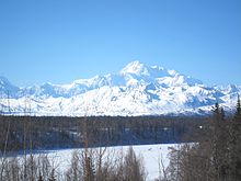 A mountain is in the background of a forest of bare trees and an icy lake.