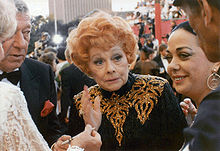 An aged Ball standing in a crowd of celebrities, wearing a black and gold sequinned dress with her characteristic red hair, looking fragile.