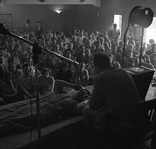 A mostly seated crowd watches as Hubbard, seated on a chair, speaks to a woman lying prone in front of him.