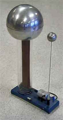 Large metal sphere suppported on a clear plastic column, inside of which a rubber belt can be seen. A smaller sphere is supported on a metal rod. Both are mounted to a baseplate, on which there is a small driving electric motor.