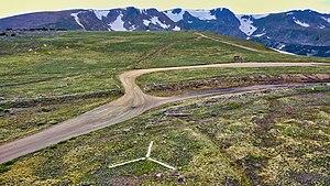 An aerial view of the emergency backcountry helicopter landing zone located at the summit of Rollins Pass; the white prong of the landing zone, furthest from the camera, points north. The comparatively low saddle of Rollins Pass is visible in this image as the summit itself (in the foreground and midground) is plainly lower in elevation than the surrounding mountains. The wood debris consists of both remnants from snowsheds that covered the tracks as well as discarded railroad ties that were removed from service in the summer of 1936.