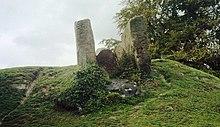 Three large stone slabs forming a chamber, as seen from below. The sky behind is white, and the stones are surrounded by green grass.