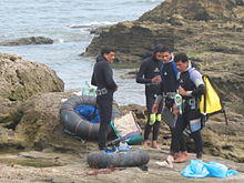 A croup of three divers dressed in wetsuits standing on a rocky shore with the sea in the background. On the ground are inflated truck inner tube floats with nets to support their catch