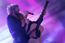 Upper body shot of Pnau performing. Nick Littlemore is shown in right profile, he is holding a microphone in his right hand and singing with his eyes closed. He wears a dark blue shirt, dark pants. At his right hip in his belt is an electronic device. Peter Mayes is partly obscured, he leans in close to Littlemore. Mayes is singing and playing a 12-string guitar with his left hand high on the fret board. His right hand holds the guitar pick as he strums the strings. His eyes are open and looking at Littlemore.