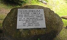 A circular boulder takes up most of the image; behind it can be seen green grass. On the boulder is a light-grey plaque. On this plaque are written the words: "Coldrum Stone Circle has been vested in The National Trust as a Memorial to Benjamin Harrison 1837–1921 of Ightham 10 July 1926". All the letters are in capital letters; those stating "Coldrum Stone Circle" are larger than the others.