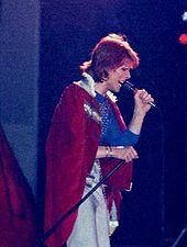 Bowie with red hair man looking to the left holding a microphone and wearing a red and white fur coat