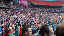 A three-tiered football stadium stand, the bottom two full of people clad mostly in white and orange. Several white and orange flags are visible.