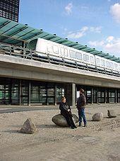 Two people look up at a train stopped at an elevated glass and concrete station