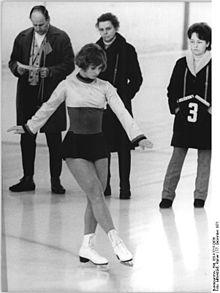 Black and white photo of a young woman, dressed in a short dress and white ice skates, executing figures on ice, while three people, two men and one woman, look on behind her