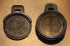 Two circular bronze plates with square handles. The plate on the right is thicker, has four characters embossed into it, and contains a rim around about three-fourths of the edge of the plate. The plate on the left is thinner, and contains a rim that, when the two plates were stacked on one another, would perfectly meet the edges of the first plate.