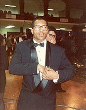 A man wearing a tuxedo and eyeglasses; other people can be seen in the background, along with folding chairs.