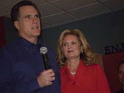 A tall, well-groomed pale-skinned man in his fifties with slightly greying dark hair wearing a dark blue shirt and holding a wireless microphone; to his side, a pale-skinned woman in her fifties with shoulder-length blond hair parted on the side, wearing red lipstick and a red jacket that almost obscures a double strand of white pearls around her neck; she is looking at him as he looks out at an unseen audience