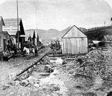 A black and white photo of a Barkerville street. On the left, a series of single-storey houses follow the road into the distance; the closest one bears a sign for the "Barnard Express." In the centre, the road edges onto a shallow ditch, flanked by rocks and mining rubble. A lone one-storey building with a sloped roof stands at the end of the ditch, with its windowless side wall facing the viewer. To the right of the photo, more single-storey structures are seen some 50 metres away.