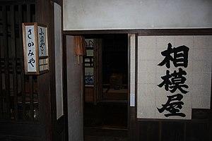 Dimly-lit narrow shopfront, with large black characters written in a single column on a shoji. A washi-and-frame lantern, mounted to protrude from the pillar, also bears signage.
