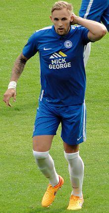 A footballer wearing a blue kit and orange boots runs with one arm raised to his head and his mouth open.
