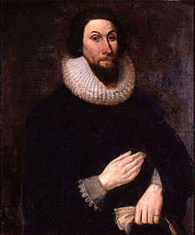 A painting of a man with a stern expression on his face, wearing very dark clothing so that his pale hands show boldly. His hands are placed in front of him, separately, one above the other.