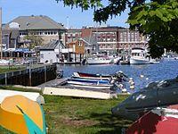 In this view of the Marine Biological Laboratory campus, the viewer stands behind rowboats resting on the grassy edge of the harbor. In the harbor, which lies in the midground, small white yachts and colorful speedboats are moored.