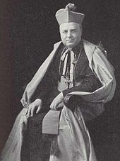 A bespectacled man holding a crosier in his left hand and wearing a mitre and liturgical vestments faces forward.