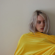 Eilish is seen wearing all yellow, leaning against a wall and looking to the side.