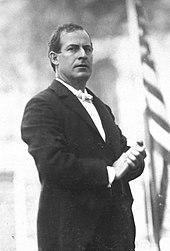 Three-quarters standing portrait of Bryan in a dark suit and white tie, with hands clasped before him, and with a serious and commanding expression