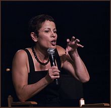 A woman with short brown hair and a black top is talking into a microphone while gesturing with her left hand.