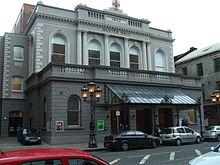 Photograph showing the front of Ulster Hall, a 19th-century grey and white rendered-brick building with black wrought-iron and glass canopy outside the front triple doors. Photo taken from the opposite side of a street, with cars parked on both sides.