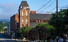 A brick tower with peaked roof on the right-hand side of a street in front of the camera. There are green hills in the background.