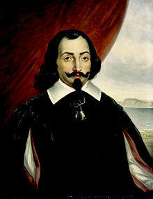 A half-length portrait of a man, set against a background that is a red curtain to the left and a landscape scene to the right. The man has medium-length dark hair, with a goatee and a wide mustache that is crooked up at the ends. He is wearing a white shirt with a wide collar, covered by a darker surcoat. There is also a bright red cape.