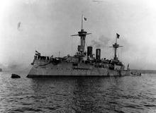 A large warship moored to a buoy, with a group of sailors crowded together on the bow