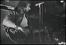 Upper body shot of a man playing a guitar with letters G-e-o visible. His head is turned partly to his left. There is a microphone stand in front of him. Other musical and studio equipment is around him. Another musician is obscured at bottom right.