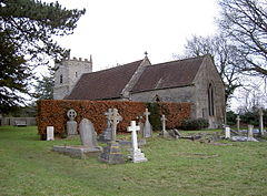 Grey stone building with red tiled roof, partially obscured by a hedge. A square tower is at the far end. The foreground includes several crosses and gravestones.