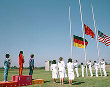 On a grassed outdoor field, three women stand on a podium to the left of the shot, while people wearing all white raise three flags on flagpoles situated at the right of shot.