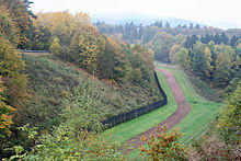 At the base of a wooded slope to the left, there is a black fence running into the distance, flanked on the right by a strip of closely cropped grass, then a strip of bare soil, then another strip of grass, and then a patrol road. Halfway up the wooded slope, a white cross is visible.