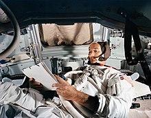 Collins lying down in the command module simulator, checklist in hand
