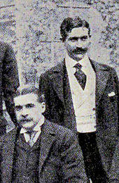 Sullivan, seated, with Herbert standing behind his left shoulder; both are very well-dressed and mustachioed