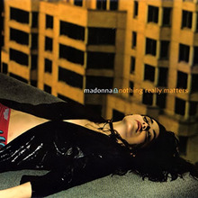 Madonna laying down at the corner of a rooftop, with yellow buildings present behind her. She wears a black shining jacket.