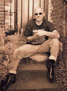 Man sitting on back door steps, wearing jeans, boots, black T-shirt and sun glasses. Rests left elbow on left knee with left forefinger pointing to his right. Right fist hidden inside left hand and he is facing forward.