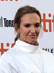 Upper body shot of a 46-year-old woman. She has a slight smile. She wears her dark hair tied at the back and is in a white top with its collar up high. Behind her is advertising for the event, which is obscured.