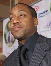 An African-American man with short hair and stubble, wearing a black suit and tie.