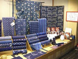A display table diaplaying a number of different indigo-dyed kasuri fabrics. A number of fabrics are displayed stacked on wooden stands in roll-form, displaying a variety of different patterns and colours ranging from the lightest blue to the deepest indigo. A small length of kasuri is displayed flat in front of the rolls; it shows a woven design of swallows or plovers. Behind the rolls of fabric, four different types of kasuri are displayed hanging on small stands, showing patterns of stripes and roundels, amongst others. At the very back, a kasuri kimono is displayed, with a small, repeating white pattern on a medium-blue base. Next to it, two large noren curtains dyed a deeper indigo, also using the kasuri technique, are displayed.
