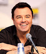 A Caucasian male with black hair and stubble, along with a black shirt. His hand is placed under his chin and a microphone is in front of him. A vague simple is marked behind him.