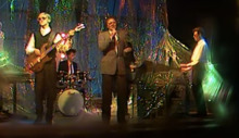 A group of people in professional attire perform in front of a sparkling curtain with lights of varying colors. The bassist (far left) and the keyboardist (middle right) wear sunglasses, while the singer (full center), drummer (center-left) and another keyboardist (far right) wear regular glasses.