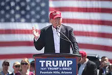 Trump speaking in front of an American flag behind a podium, wearing a black suit and red hat. The podium sports a blue "TRUMP" sign.
