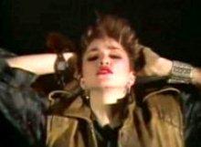 A brunette Madonna holding her hands behind her head. Her garments include a khaki shirt and sleeveless jacket.