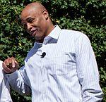 A man wearing a buttoned shirt stands in front of a vine and smiles.