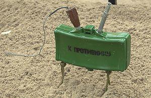 A green plastic-body mine supported by a pair of scissor legs, with "К ПРОТИВНИКУ" (K PROTIVNIKU; to the enemy) stenciled on the front.