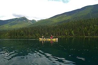 Two paddlers sit in a canoe, gently paddling across calm water. Behind them, large green mountains flank the water.