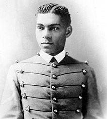 Cadet Henry O. Flipper in his West Point cadet uniform. It has three larger round brass buttons left, middle and right showing five rows. The buttons are interconnected left to right and vice versa by decorative thread. He is wearing a starched white collar and no tie. He is a lighter-colored African American with plated corn rows of neatly done hair. He is facing the camera and looking to the left of the viewer.