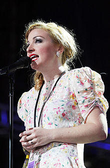 A 29-year-old woman is singing into a microphone while shown in a three-quarter shot. She is slightly turned to her right and is staring forwards. She wears a floral, summery dress and holds her hands clasped at her waist. She also wears a lanyard with black cord over her neck and another thicker and grey one on her right wrist. A gold ring is on her left hand. Her blonde to brown hair is tucked to her right shoulder.